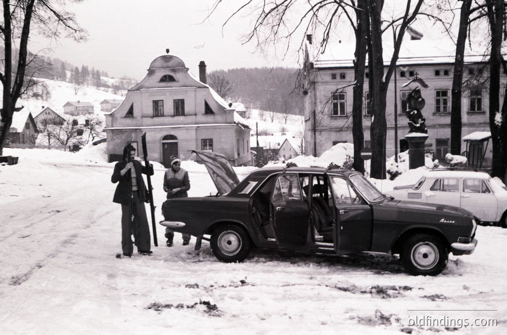Vintage black-and-white street scene in a European town during winter, featuring a 1960s-era Zastava 750 car with open hood. Two men in winter coats pose near the vehicle, surrounded by snow-covered rooftops and parked cars. Architectural details include a distinctive gabled building and a lamppost.