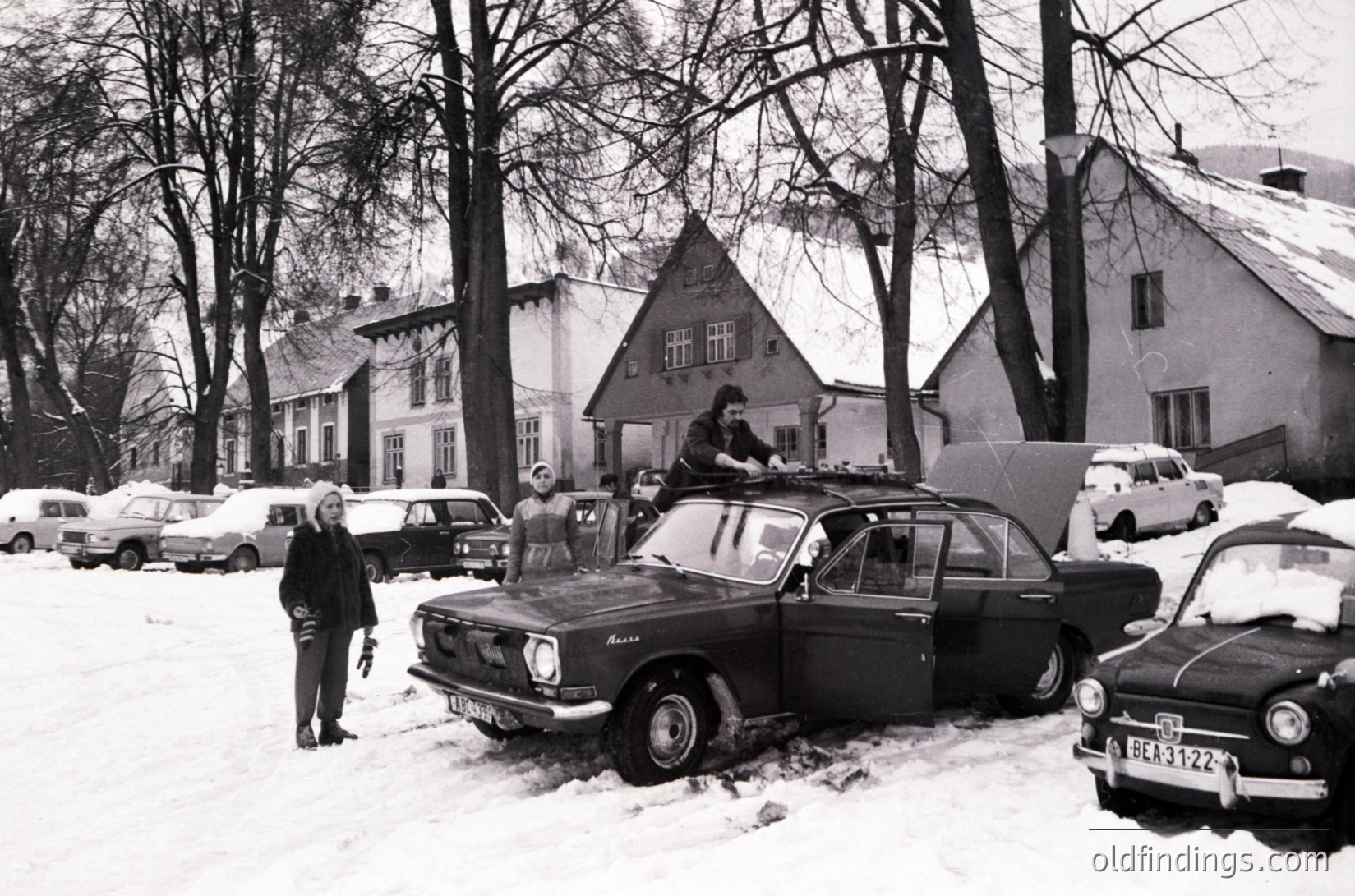 Vintage black-and-white street scene in a snowy European neighborhood, featuring mid-century cars (including a Zastava/Karola) and classic vehicles. People in winter coats gather around parked cars, with a house and trees in the background. Likely Eastern Bloc, 1960s-1970s.