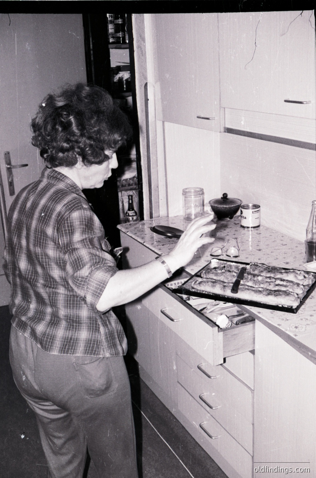 Mid-century kitchen scene featuring a person in a plaid shirt preparing food on a retro stove, 1960s-1970s. Wooden cabinets, vintage jars, and a manual clock radio on the counter. Classic domestic interior with utilitarian design.