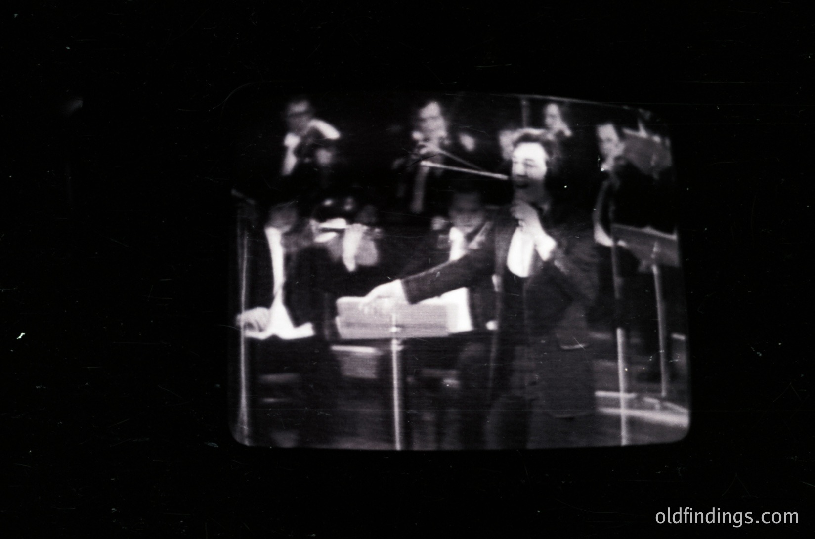 Vintage black-and-white photo of a conductor leading an orchestra, likely mid-20th century. Formal attire, sheet music, and baton suggest a classical performance. Stage lighting and blurred audience indicate a concert hall setting.