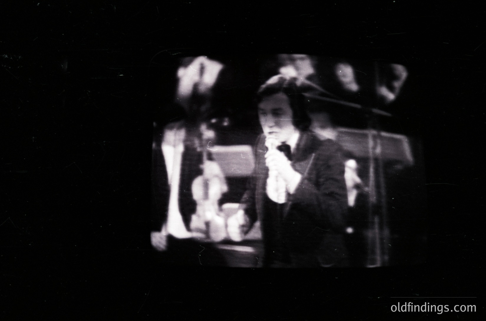 Vintage black-and-white photo of a silent film scene. A performer in a fedora and suit gestures dramatically while holding a prop resembling a small, ornate box. Stage lighting and shadowy background suggest early 20th-century cinema ( ).