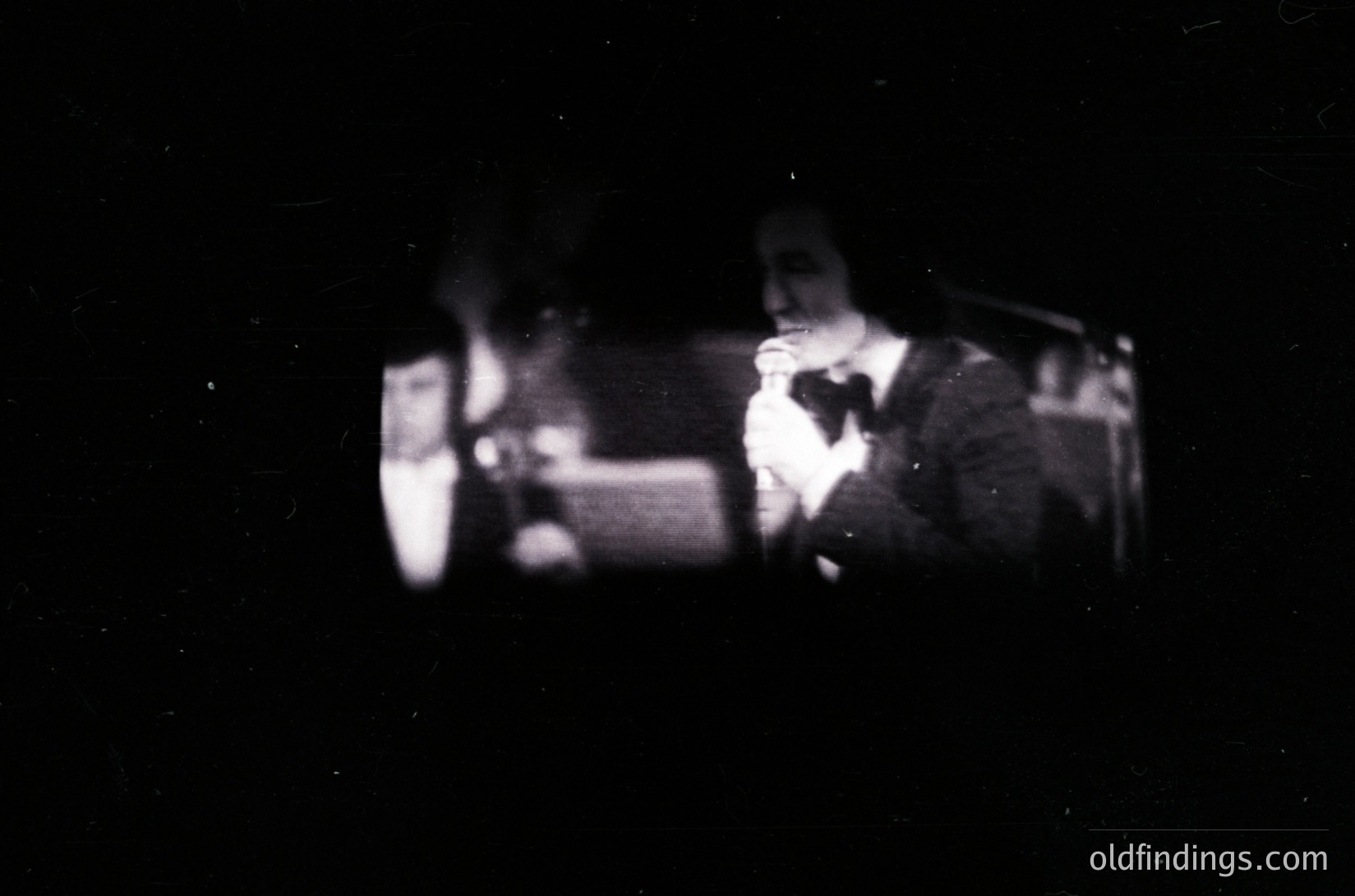 Vintage black-and-white photo of two musicians performing live, likely in a dimly lit venue. One plays guitar while the other sings into a microphone. Artistic grain and low contrast suggest mid-20th century (1950s–1970s) photography. Ideal for vintage music, indie film, or retro aesthetic projects.