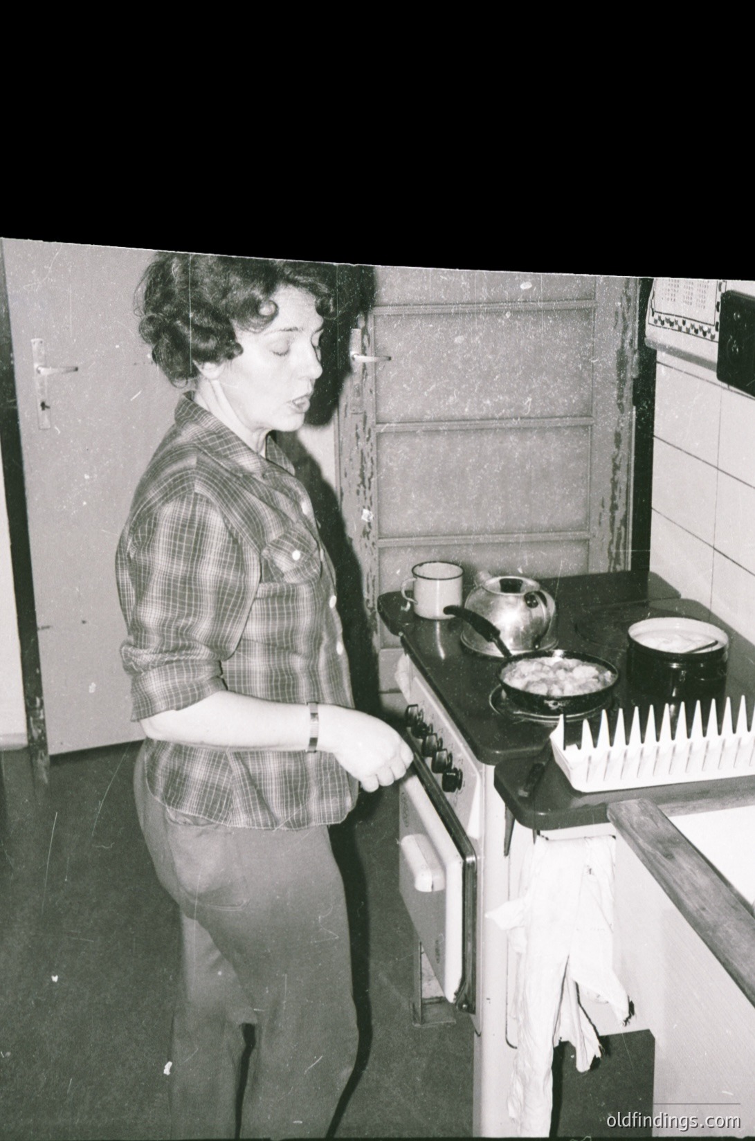 A woman in a plaid shirt and rolled-up trousers operates a vintage gas stove, cooking in a mid-century kitchen. Two mugs and a pot sit on the counter; a towel hangs nearby. Likely – domestic scene, reflecting post-war kitchen design.