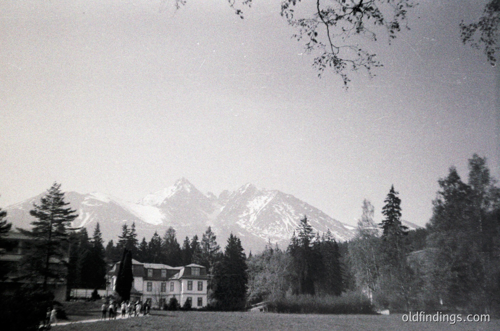 Mid-century alpine resort building nestled in a forested valley, framed by snow-capped peaks. Classic timber-framed architecture with large windows, likely 1950s-60s.
