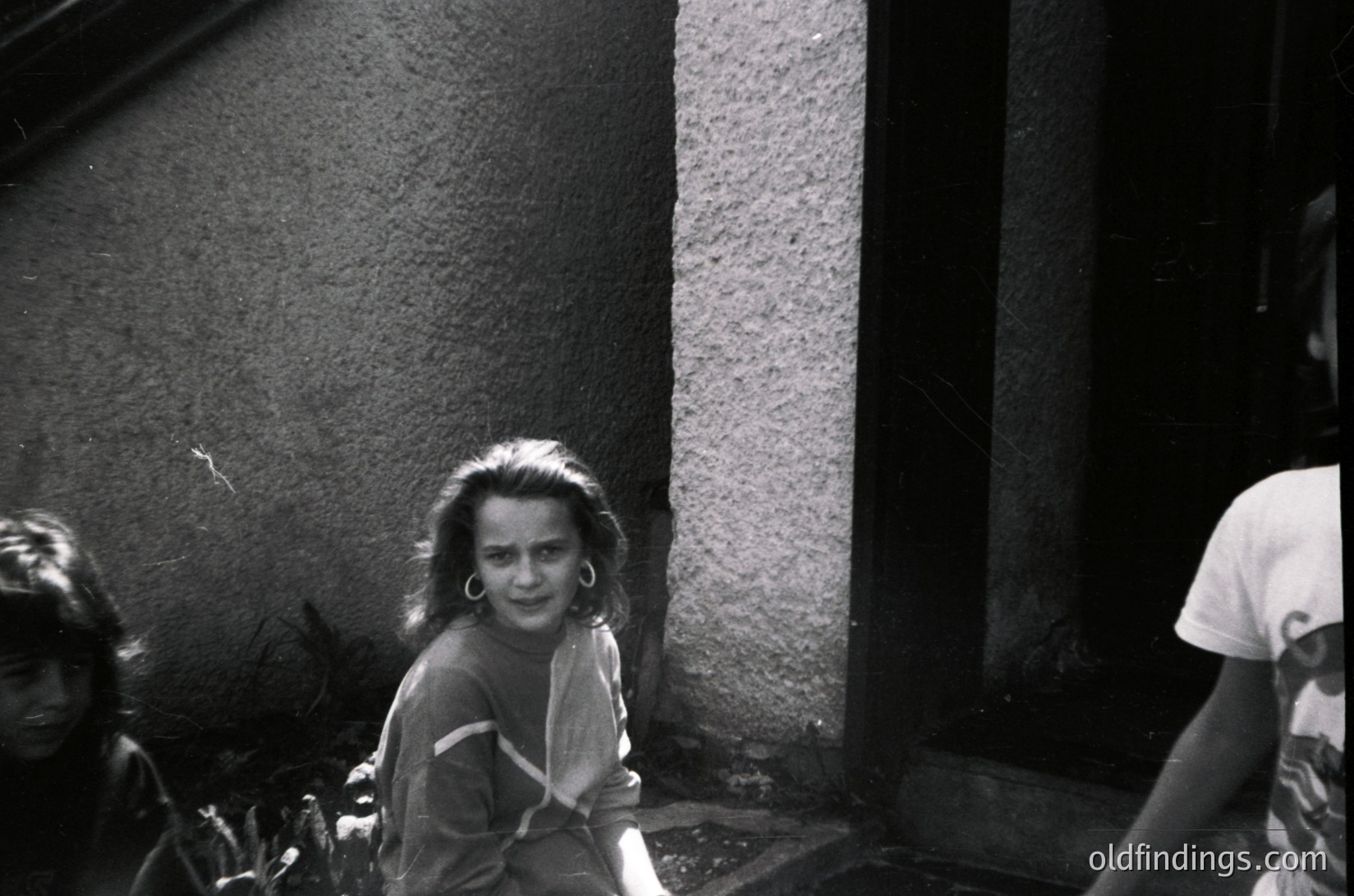 Vintage black-and-white candid of three children in an urban alleyway, likely 1960s–1970s. Girl in center wears layered clothing and hoop earrings; others partially visible. Rough concrete walls and worn pavement suggest post-war European setting. Candid, unposed street photography captures youthful energy.