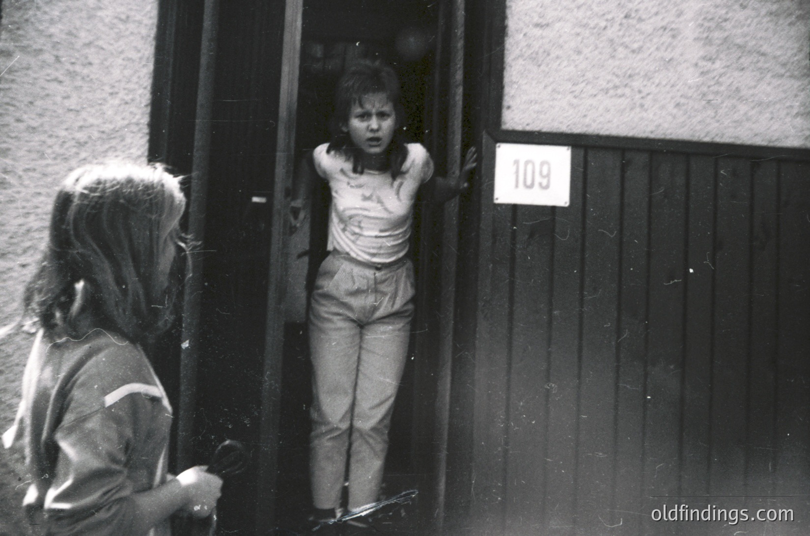 Two children in mid-20th-century streetwear—one leaning on a gate, the other peering through an open door marked "109"—capture candid childhood curiosity. Concrete block building suggests urban residential setting. Black-and-white grain hints at 1950s–1970s era.