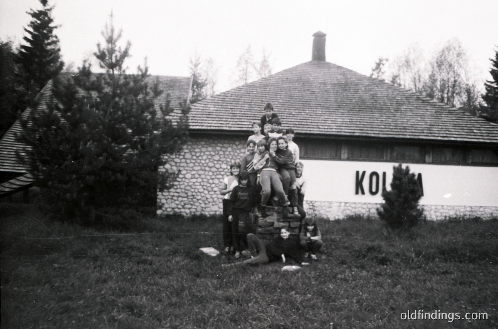 Mid-20th century black-and-white photo: Group of 12+ children stacked atop wooden crates in front of a rustic stone building labeled "KOLHOZ." Rural setting with pine trees and grassy foreground.