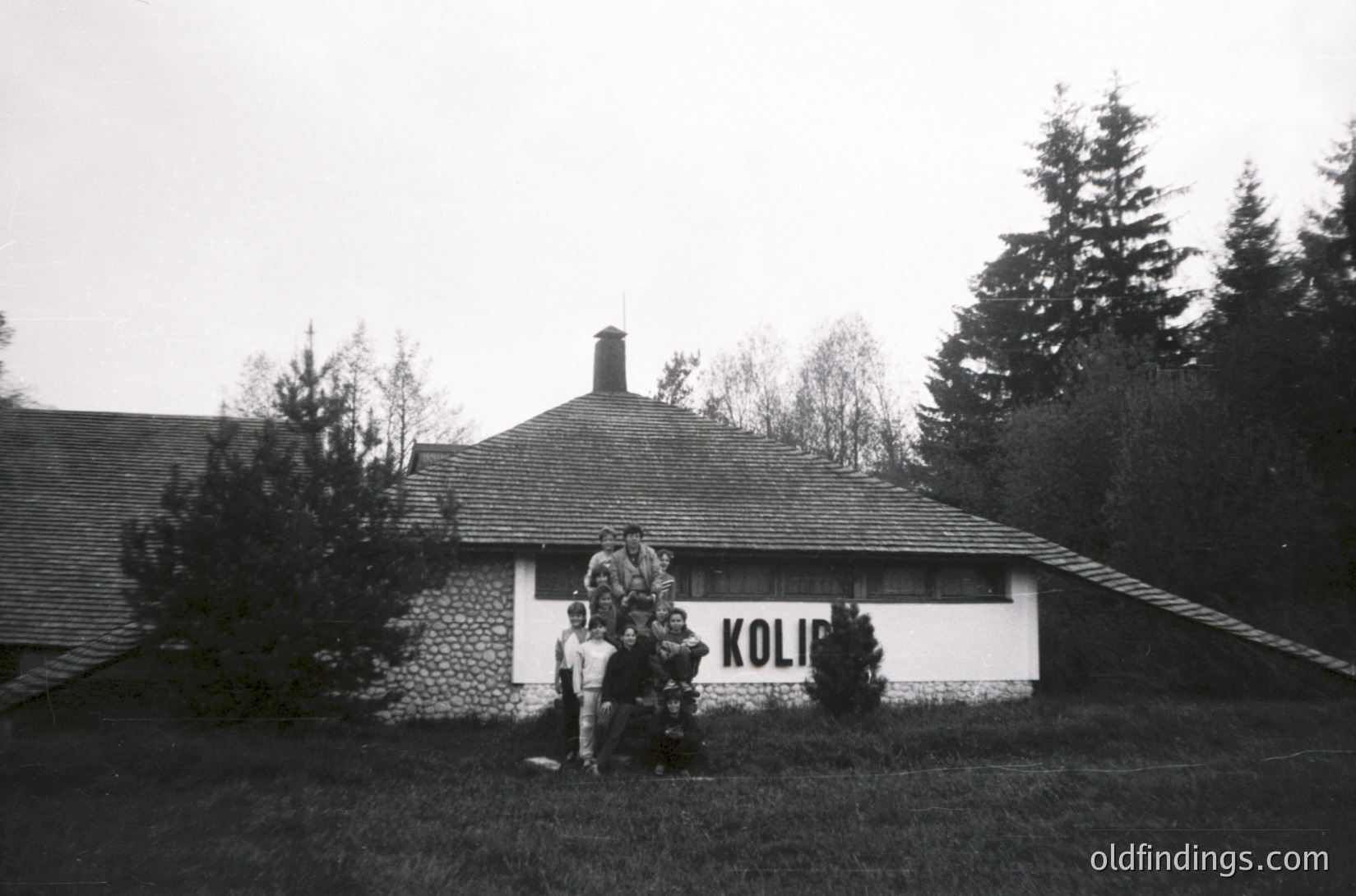Black-and-white photo of a group posing outside a rustic stone building labeled **"KOLIB"** in the 1960s–70s. The structure features a pitched roof, chimney, and a small porch. Lush evergreen trees frame the scene, suggesting a mountainous or alpine setting. The group’s attire hints at mid-century outdoor recreation.