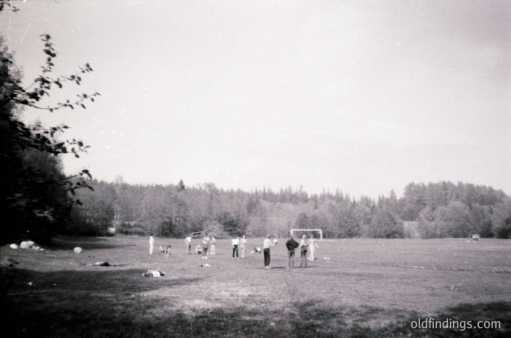 Vintage black-and-white photo of a mid-20th-century outdoor soccer game in a grassy field. Players in casual 1950s-60s attire, including knee-high socks and short pants, engage in a match near a goalpost. Dense forest and rolling hills in background.