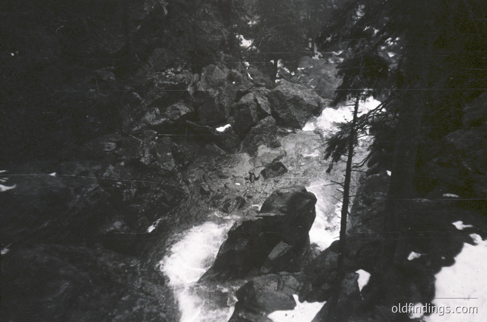 Black-and-white aerial view of a densely packed urban area with grid-like streets and industrial structures. Shadows and light contrast highlight rooftops and pathways, suggesting early 20th-century city planning. Potential European setting due to architectural style.