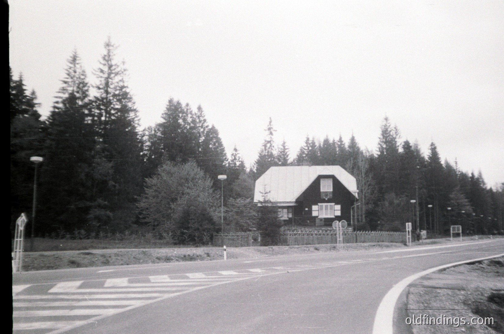 Mid-century modern house with gabled roof and white upper walls, nestled in a forested area. Concrete road curves left with pedestrian crosswalk. Streetlights and gated entrance suggest suburban or rural residential design. Likely or architecture.