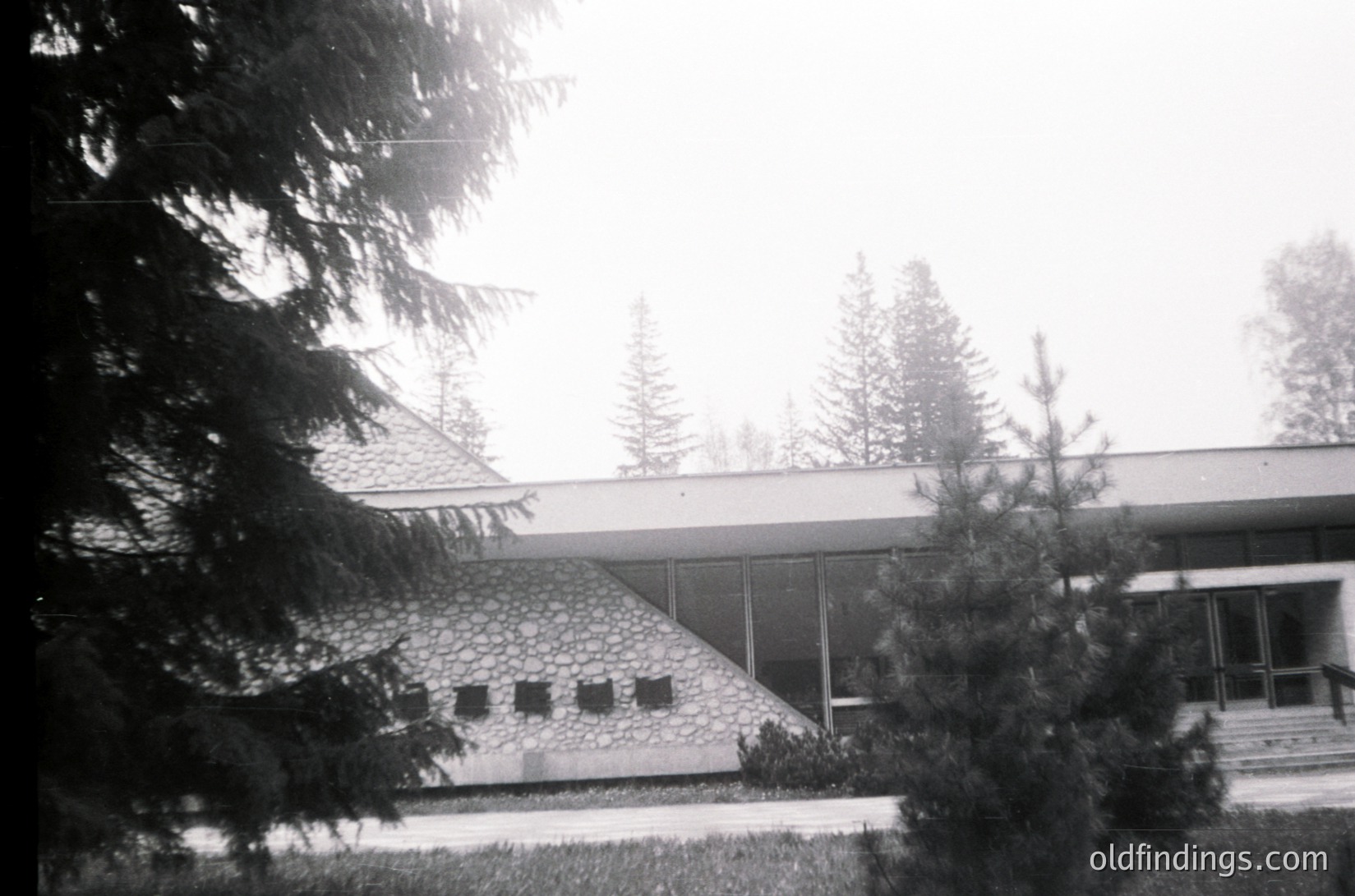 Mid-century modern building with flat roof and stone facade, framed by evergreen trees. Overcast sky suggests cloudy weather. Likely institutional or residential architecture from the 1950s–1970s.