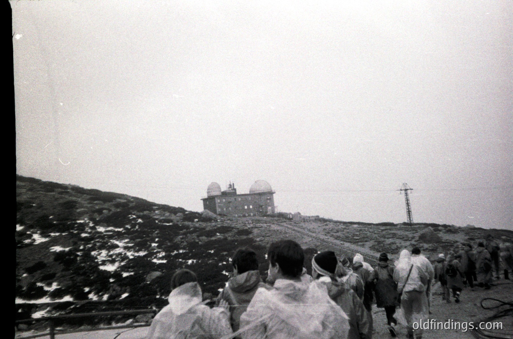 Vintage black-and-white photo of a group ascending a rocky hillside toward a pair of cylindrical observation towers. Mid-20th century attire suggests a public outing or excursion. Overcast skies and rugged terrain hint at a mountainous or hilly location, possibly a historic lookout or resort.