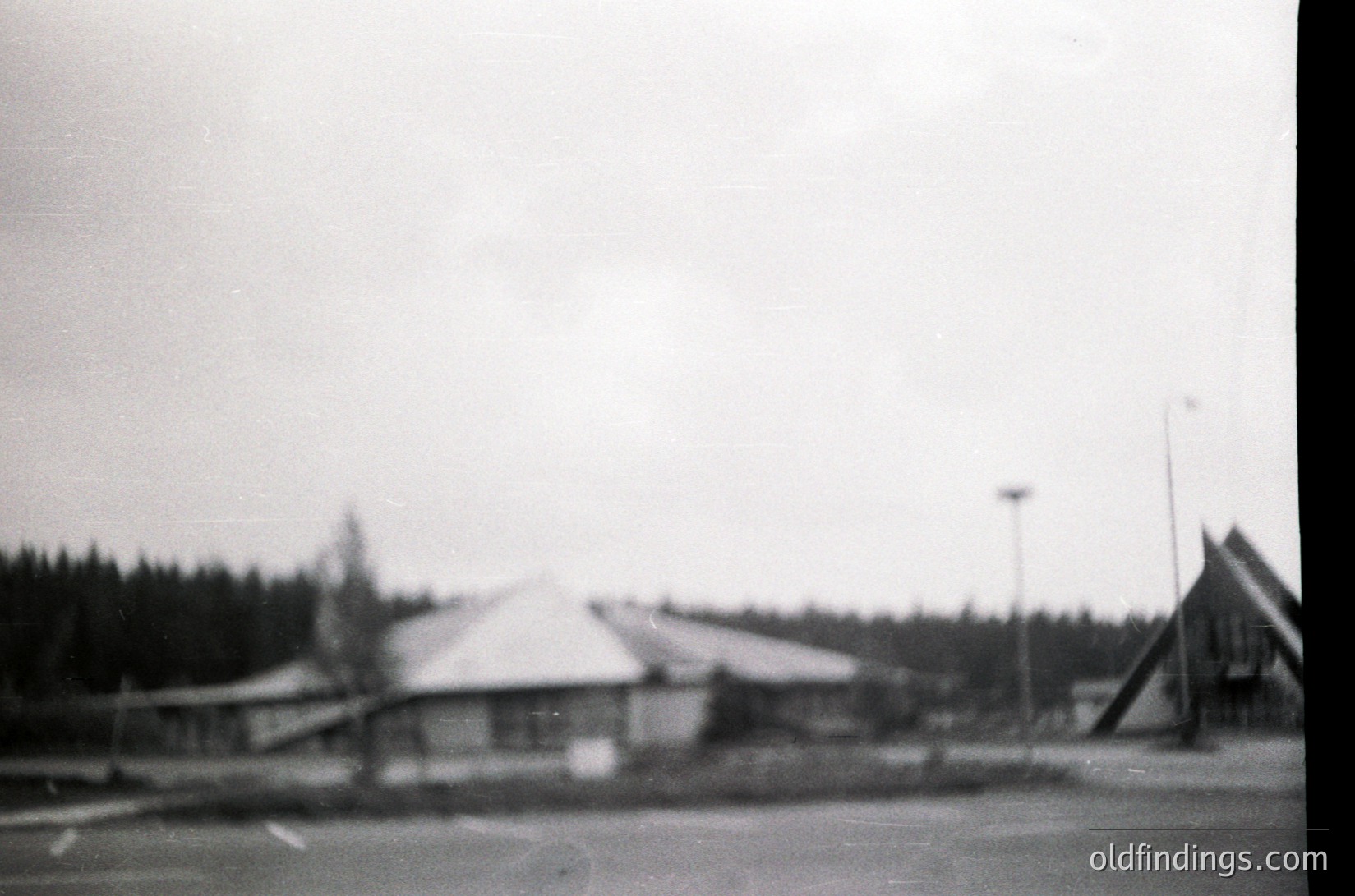Vintage black-and-white shot of a modest, single-story building with a peaked roof in a forested area. Likely a rural or suburban setting with sparse infrastructure. Visible streetlight and parked vehicles suggest a small town or village. Style hints at mid-20th century (1950s–1960s).