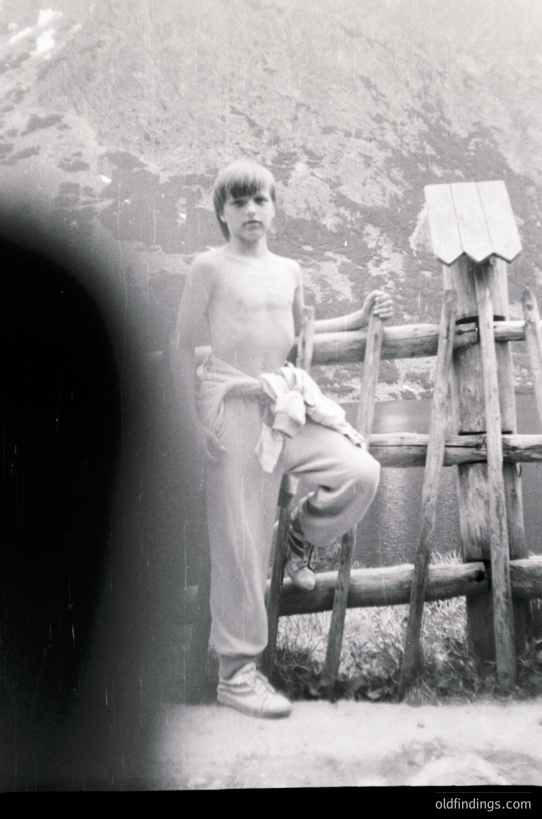 Young boy in mid-20th-century outdoor setting, shirtless with rolled-up pants, leaning on rustic wooden fence. Blurred foreground suggests vintage camera lens.