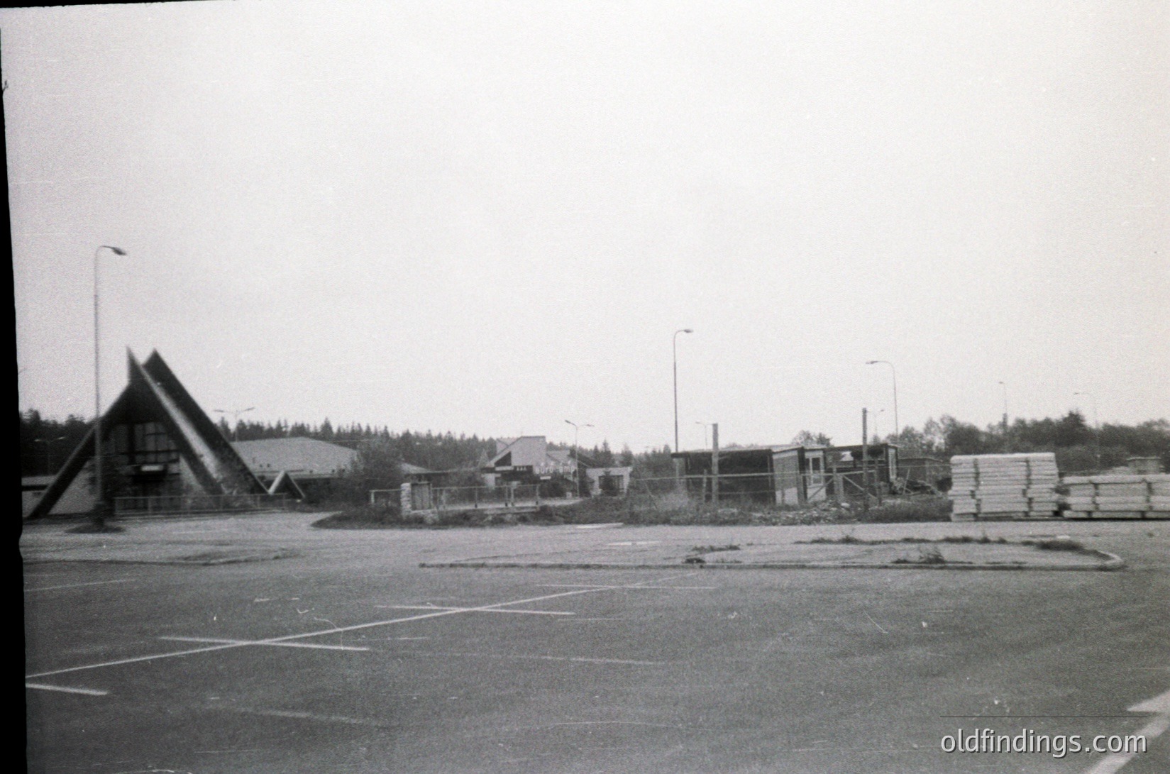 Black-and-white shot of a mid-20th-century industrial/commercial site. Prominent triangular-roofed warehouse or factory building with stacked wooden planks and construction materials in foreground. Overcast sky and sparse trees suggest a northern or temperate climate. Likely 1950s–1970s era.
