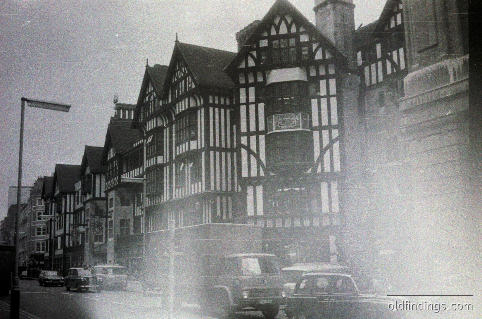 Black-and-white street scene featuring Tudor-style timber-framed buildings with exposed beams and decorative brickwork, likely mid-20th century. Classic vintage cars, including a bus and sedans, navigate a narrow road lined with street lamps. Fog or mist obscures distant details, adding atmospheric depth.