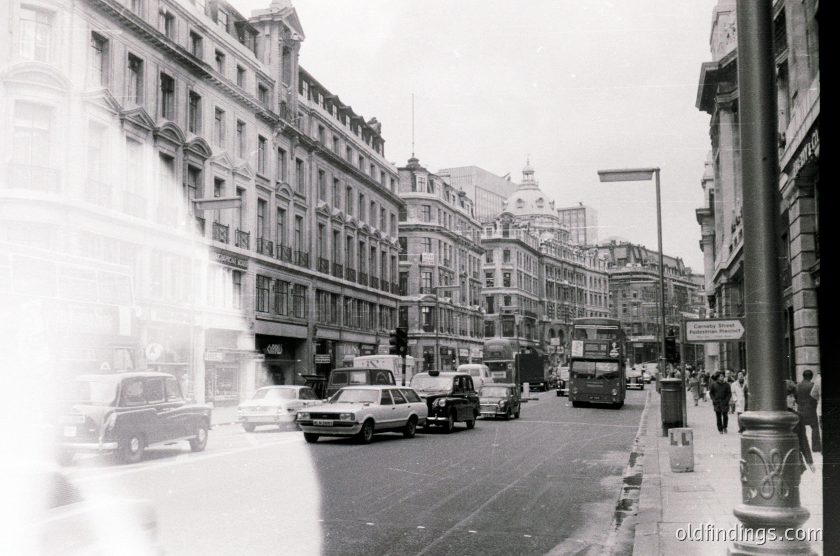 Classic mid-20th century urban street scene featuring Edwardian-era architecture. Multi-story brick buildings with ornate facades line both sides, housing commercial establishments. Vehicles—including vintage cars and a double-decker bus—traffic along a wide road with minimal pedestrians. Overcast sky enhances vintage atmosphere.