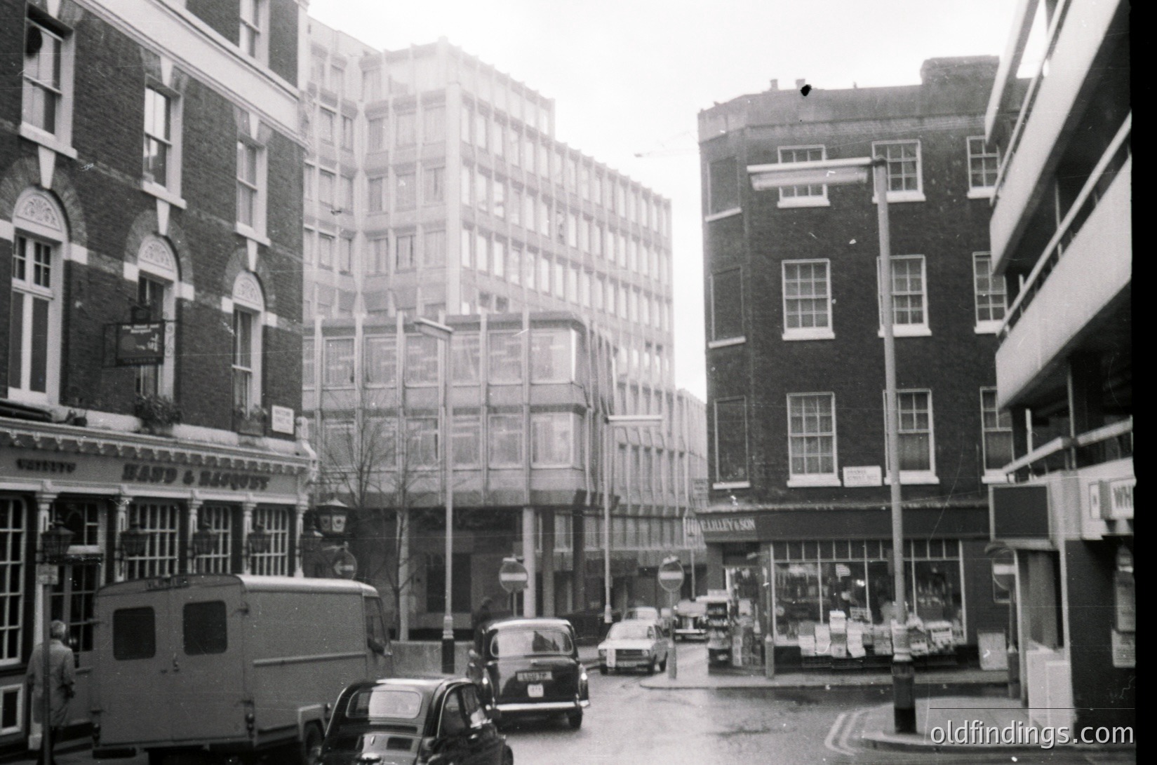 Mid-century urban street scene featuring brick facades, vintage cars, and a bus. Prominent signage reads "Lloyds Bank" and "Savoy" on corner buildings. Overcast skies and wet pavement suggest early 1960s London.