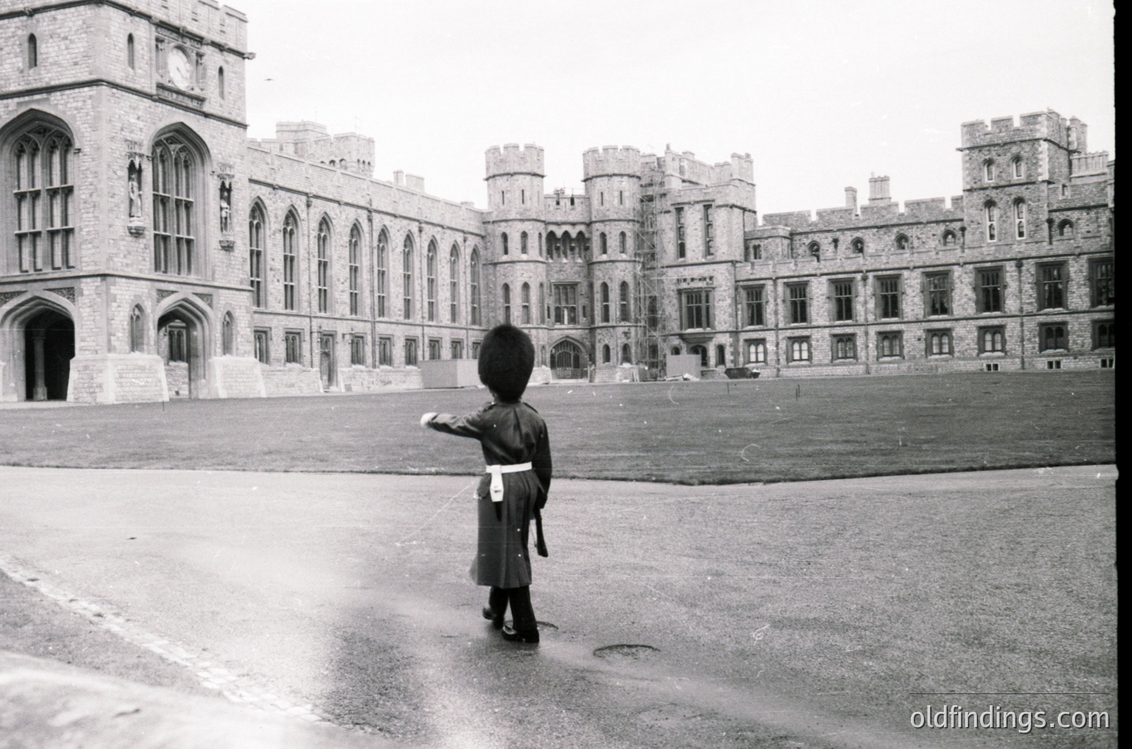 Black-and-white shot of a sentry in traditional British military uniform (helmet, sash, boots) standing at attention outside Buckingham Palace, 1950s-1960s. Gothic Revival architecture with crenellated roofline and long windows dominates the background.