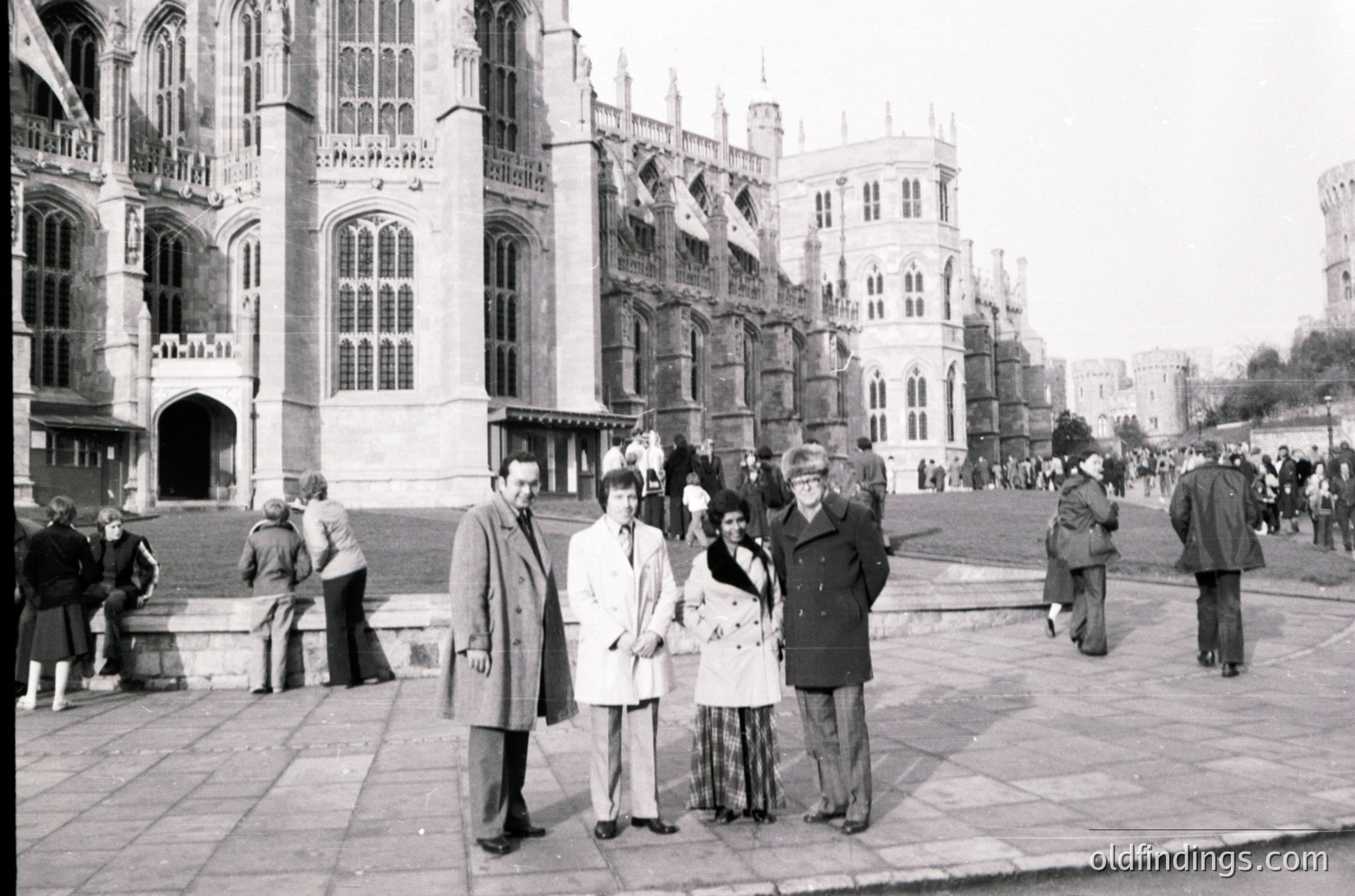 Black-and-white photo of Buckingham Palace exterior, mid-20th century. Gothic Revival architecture with pointed arches and ornate stonework dominates the scene. Six individuals pose in formal attire—men in suits, women in dresses—suggesting a royal or official event. Crowd of onlookers fills the courtyard, indicating public interest.