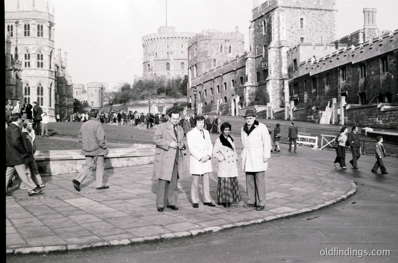 Black-and-white street scene at **Varna’s 1960s seaside fortress**, featuring medieval-style towers and fortified walls. Group of five people in mid-century attire (coats, hats) poses near a curved pavement, while others walk along the promenade. Architectural details include arched windows and a bridge in the background.