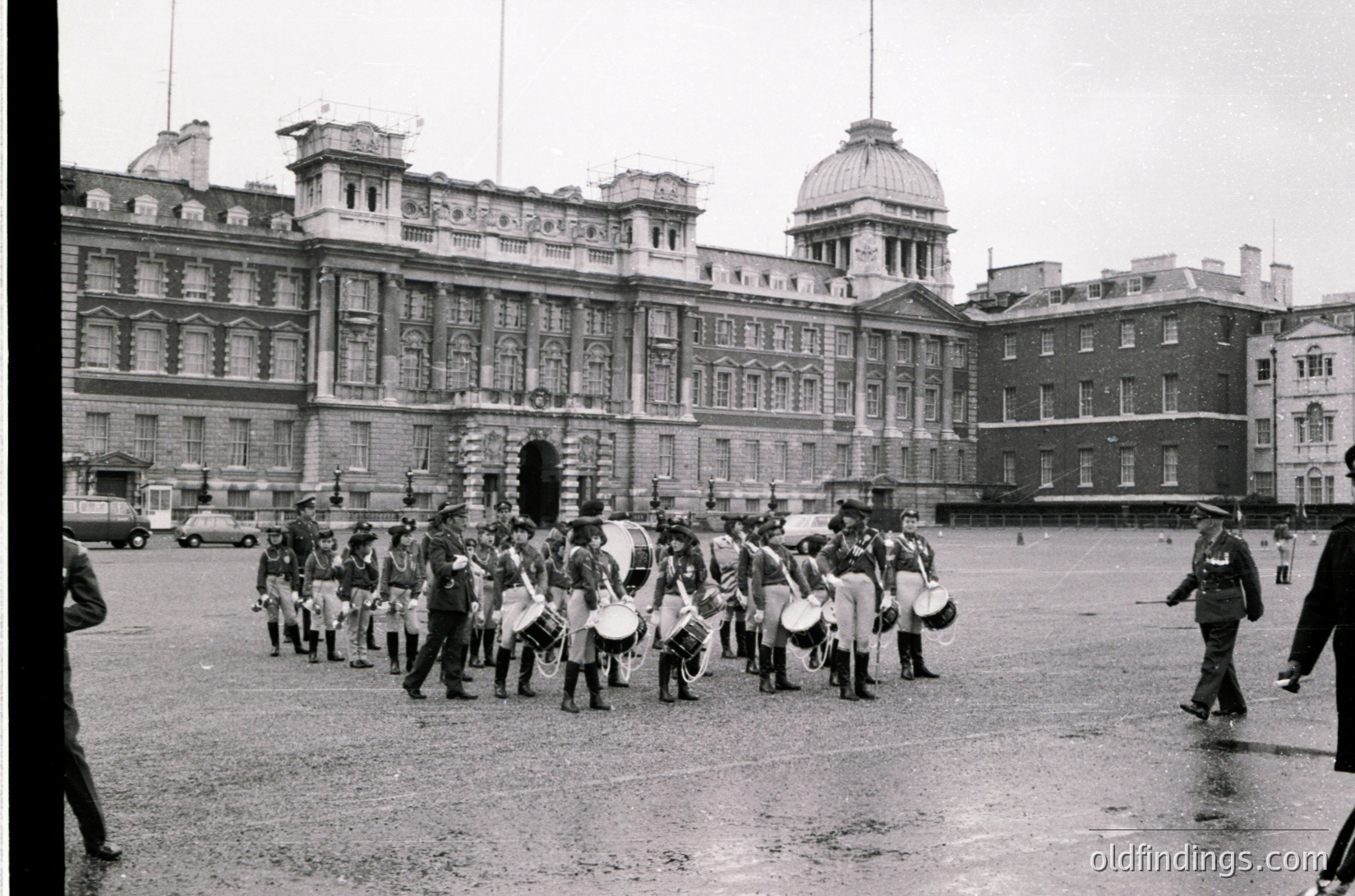Military band performing in front of Buckingham Palace courtyard, mid-20th century. Uniformed musicians in white tunics and dark trousers play drums and brass instruments. Classic neoclassical architecture with domed roof and ornate detailing visible.