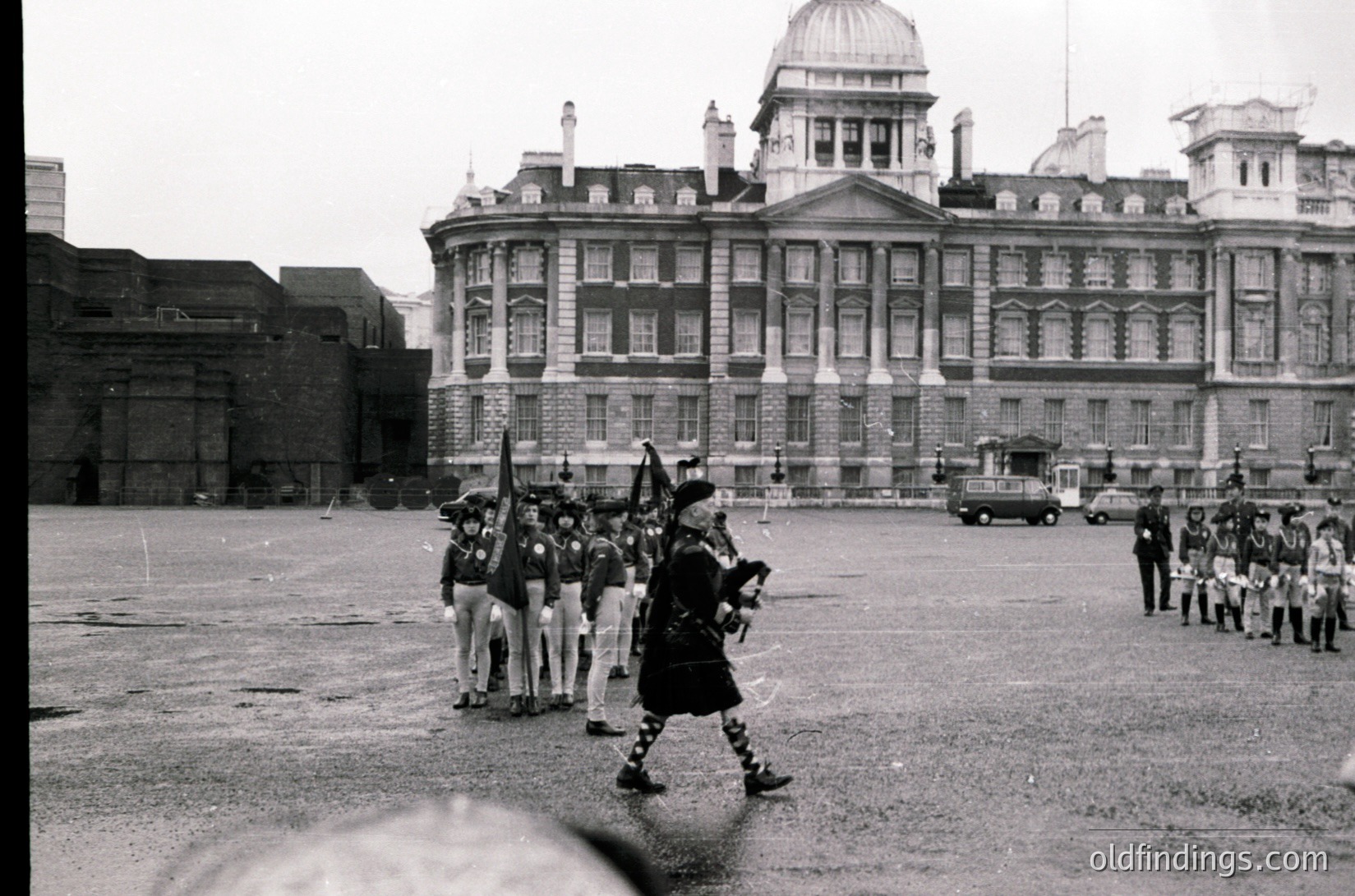Neoclassical government building with symmetrical columns and domed roof, flanked by uniformed guards in traditional kilts and hats. Mid-20th century military parade in an open courtyard.