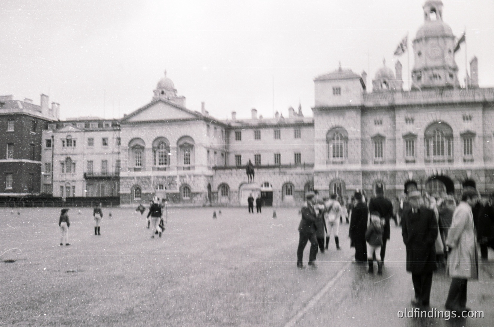Neoclassical courtyard with grand, symmetrical buildings featuring arched windows and domed roofs. Mid-20th century street scene shows men in suits and children playing soccer on wet pavement. Likely institutional or governmental complex.