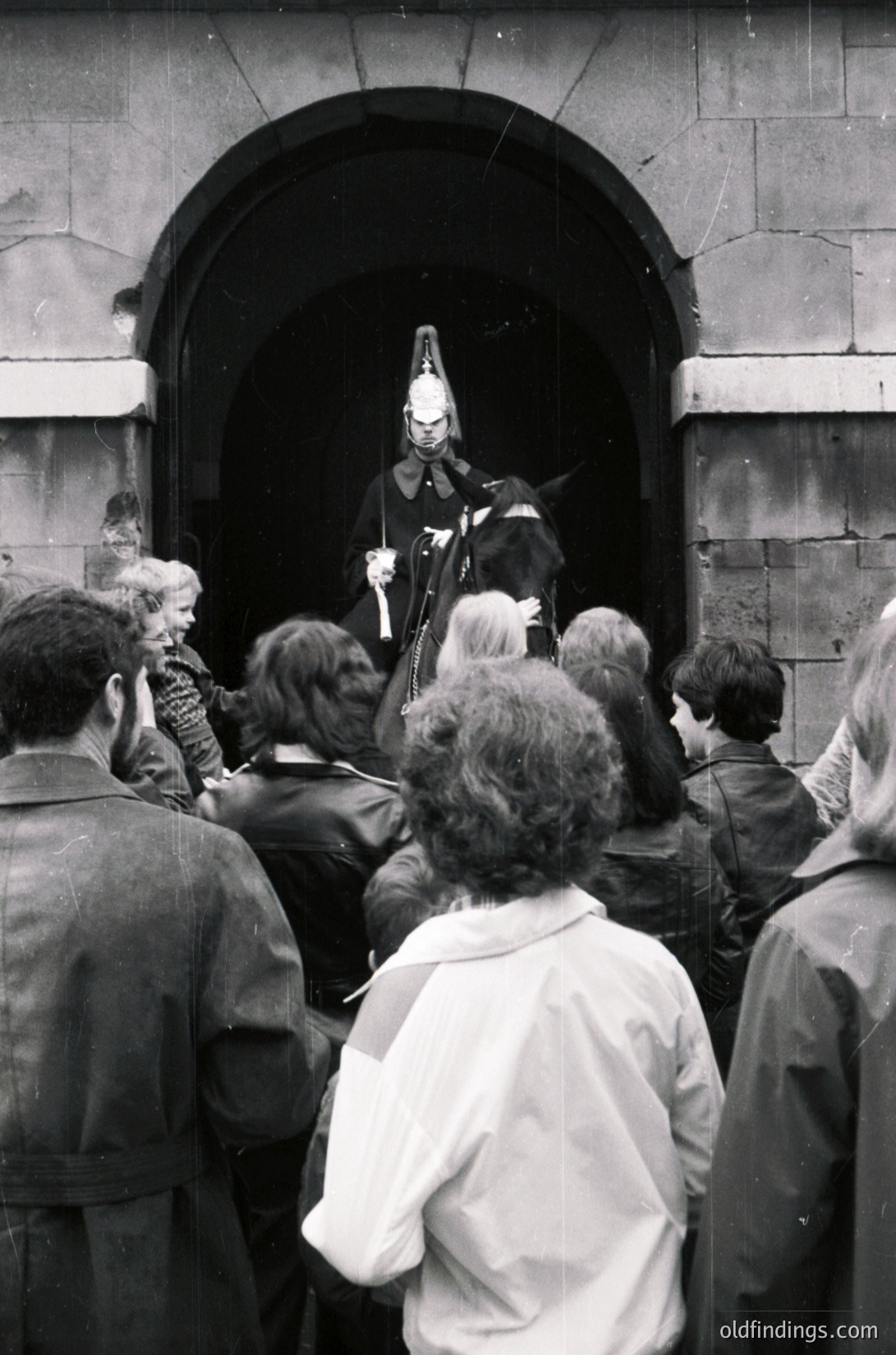 Black-and-white photo of a public event featuring a mounted police officer addressing a crowd through an archway. Uniform includes helmet and long coat; crowd wears 1960s–70s attire. Likely UK due to uniform style.