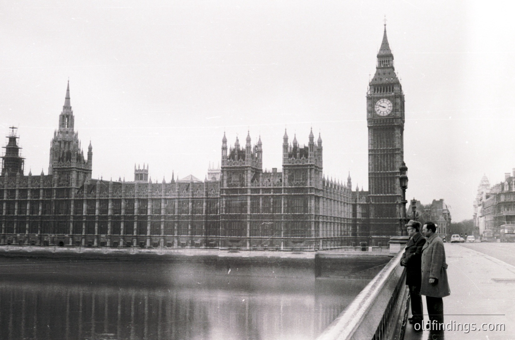 Black-and-white shot of **Big Ben and Houses of Parliament** from Westminster Bridge, featuring scaffolding on the clock tower and Gothic Revival architecture. Two men in mid-20th-century attire stand on the bridge railing, overlooking the Thames. Foggy, overcast London atmosphere.