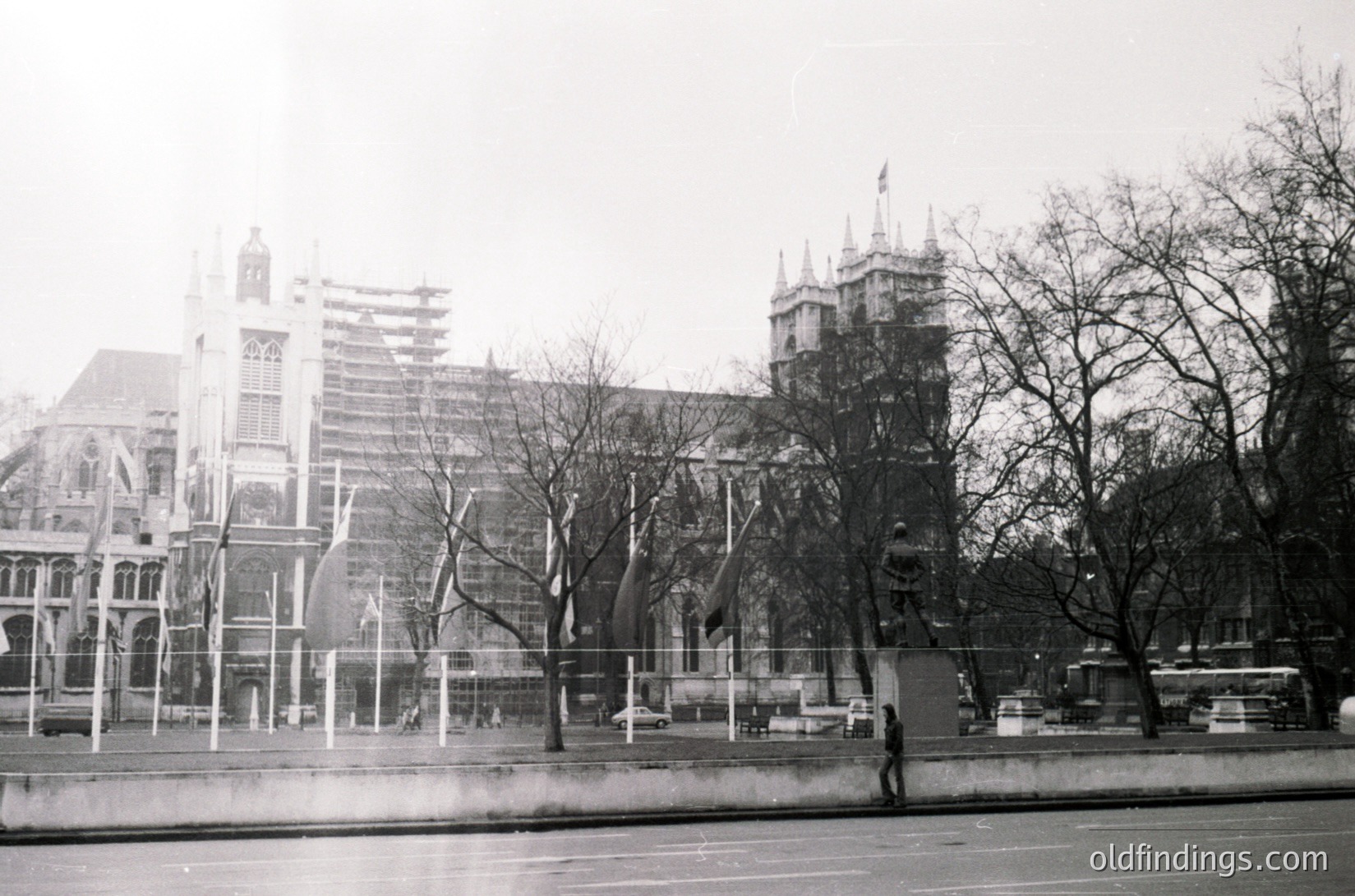 Historic Gothic Revival architecture with pointed spires and arched windows, likely St. Martin-in-the-Fields, London. Barren winter trees frame the scene, with a lone pedestrian crossing the street. Black-and-white photo suggests 1950s–1970s era.
