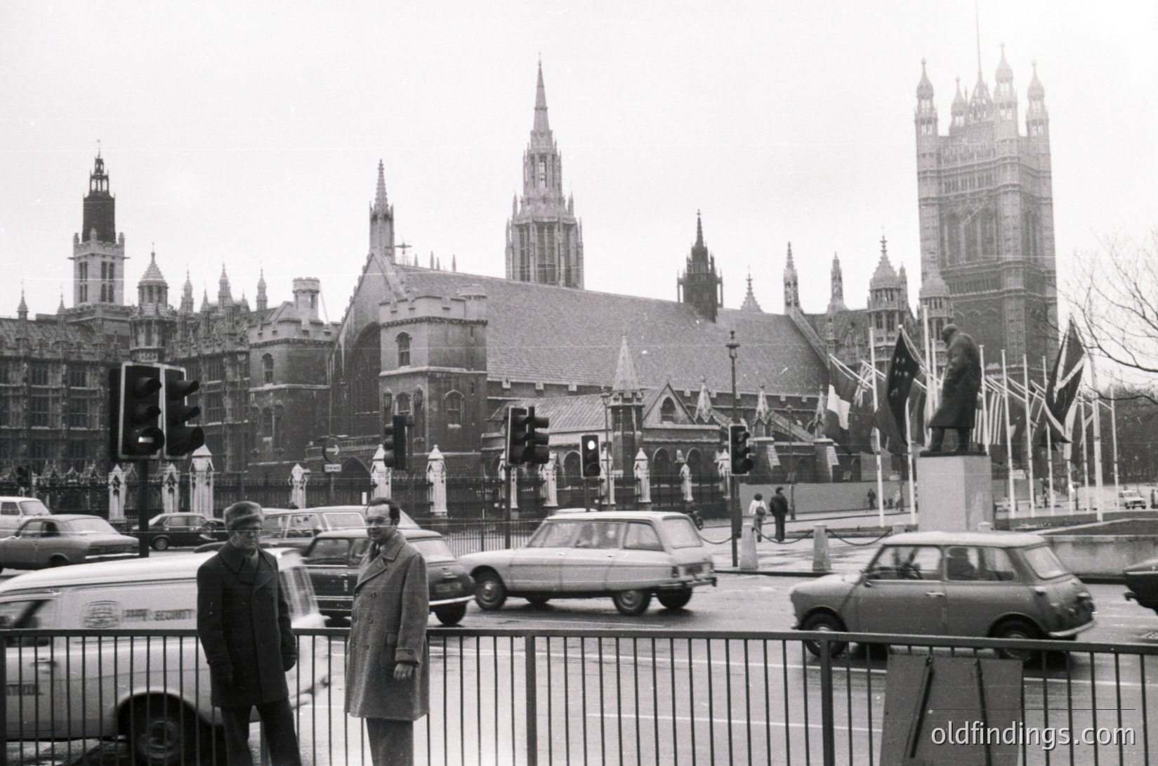 Black-and-white street scene at Westminster, London, featuring iconic Gothic architecture of the Palace of Westminster and Big Ben. Mid-20th century (1950s–1960s) vintage cars and pedestrians in classic attire. Prominent traffic lights and Union Jack flags.