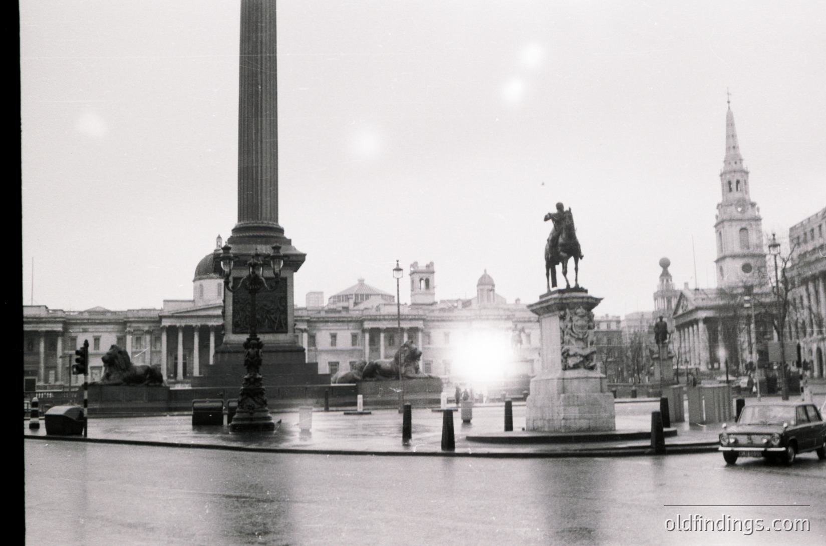 Classic black-and-white shot of Trafalgar Square’s Nelson’s Column (1840s) and equestrian statue (Charles I, 1633) in London, UK. Neoclassical architecture and fountains frame the scene. Overcast skies and wet pavement suggest early 20th-century urban photography.