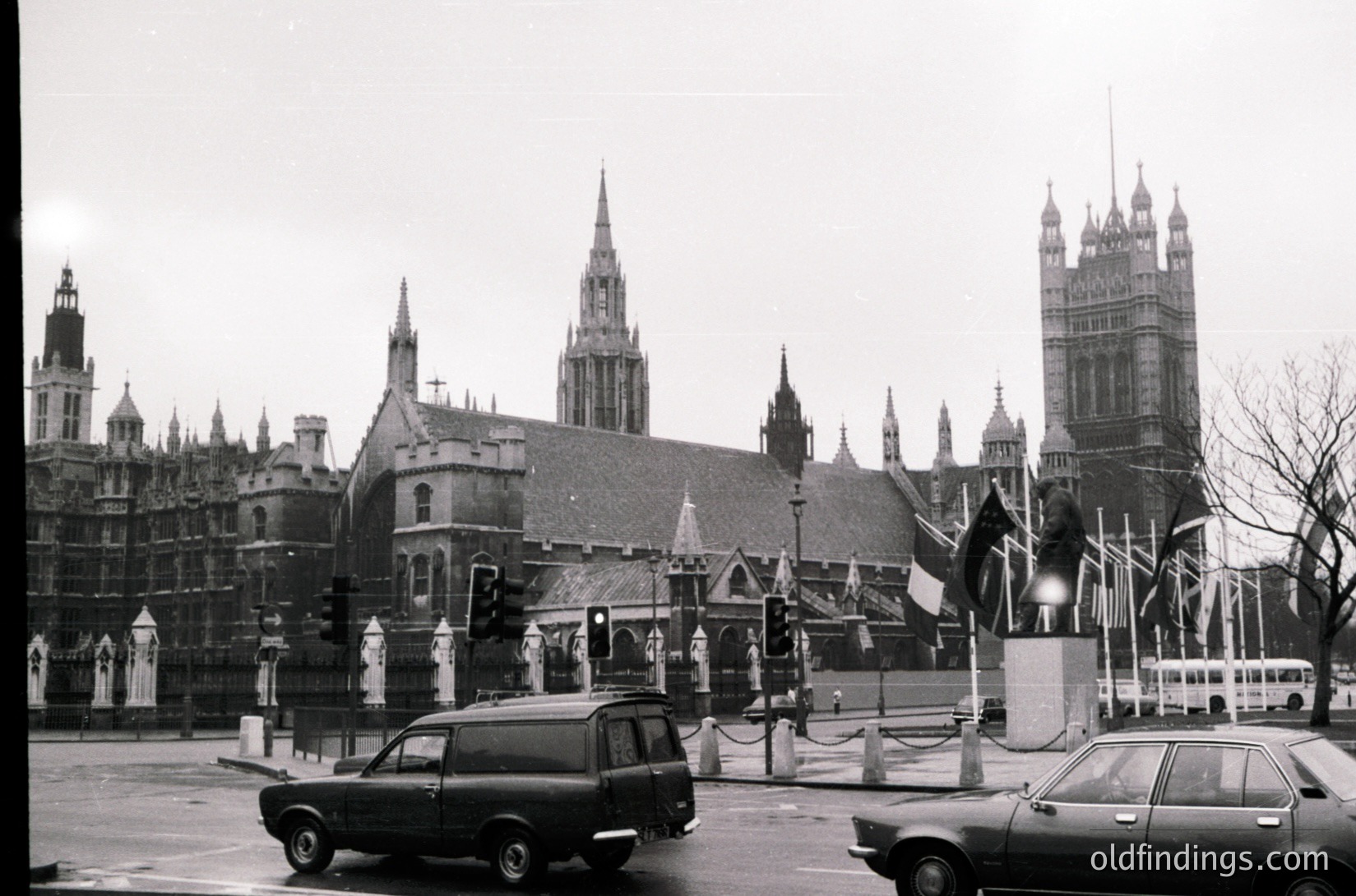 Black-and-white shot of **Westminster Abbey** and **Houses of Parliament** (Big Ben) in London, UK, c. 1960s–1970s. Gothic architecture dominates with pointed spires, flying buttresses, and intricate stonework. Vintage cars (including a station wagon and double-decker bus) navigate the street, while Union Jacks flutter on poles. Overcast sky enhances historic atmosphere.