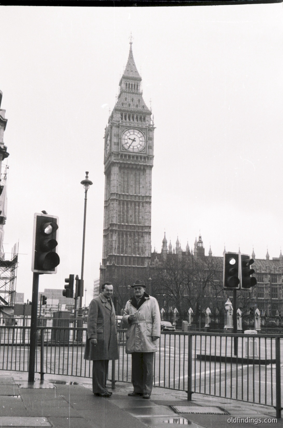 Two men in 1960s-era overcoats pose near Big Ben, London. Urban scene with traffic lights and railings. Classic black-and-white street photography.