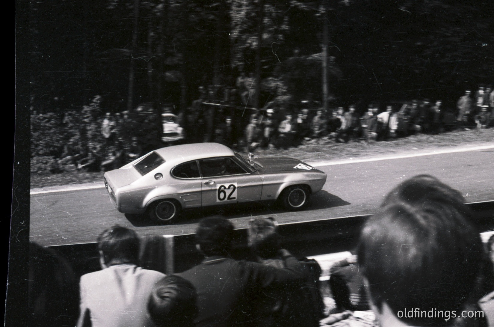 Vintage race car () in motion on a tree-lined road, likely a 1960s rally event. Crowd watches from roadside, suggesting public spectator access. Classic coupe design with minimalist livery—ideal for automotive history research.