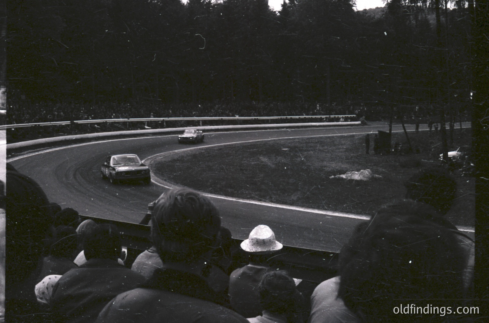 Black-and-white race scene: spectators in hats watch a vintage sports car navigate a sharp curve on a wet track, surrounded by dense forest. Mid-20th century motorsport atmosphere.