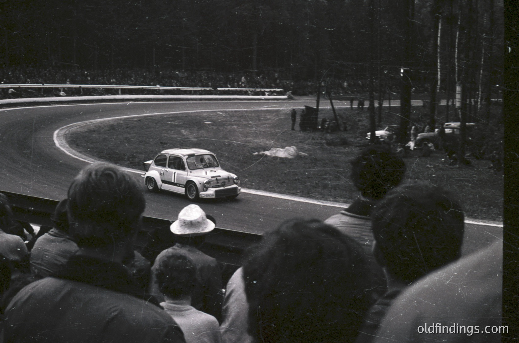 Classic 1960s rally car navigating a sharp curve on a tree-lined track, surrounded by spectators. The compact vehicle features racing stripes and sponsor decals, suggesting a competitive event. Crowds in casual attire watch from the side, indicating public interest.