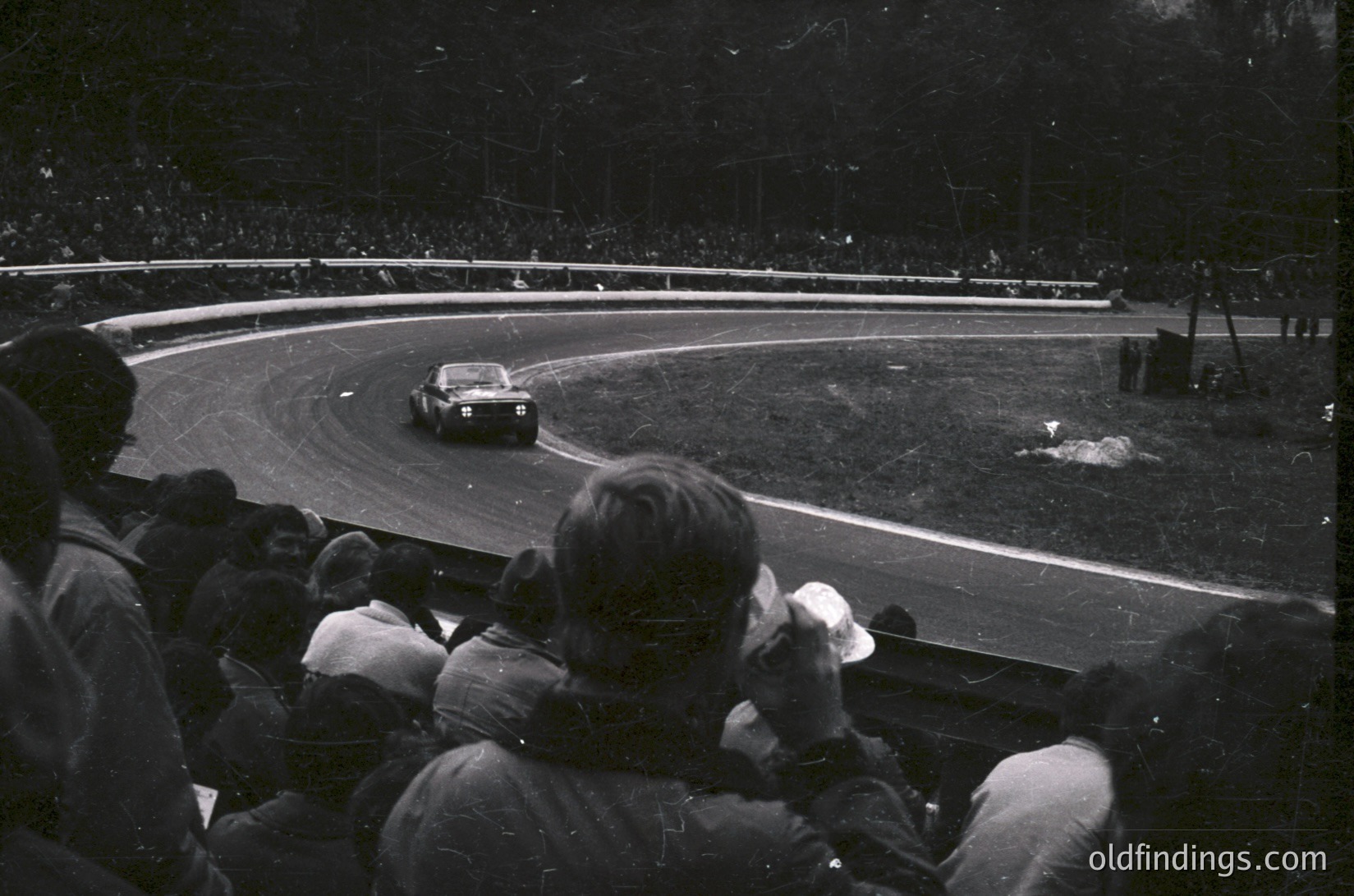 Black-and-white shot of a vintage race car navigating a sharp curve on a wet track, surrounded by spectators in grandstands. Mid-20th century motorsport atmosphere with blurred motion and dramatic lighting.