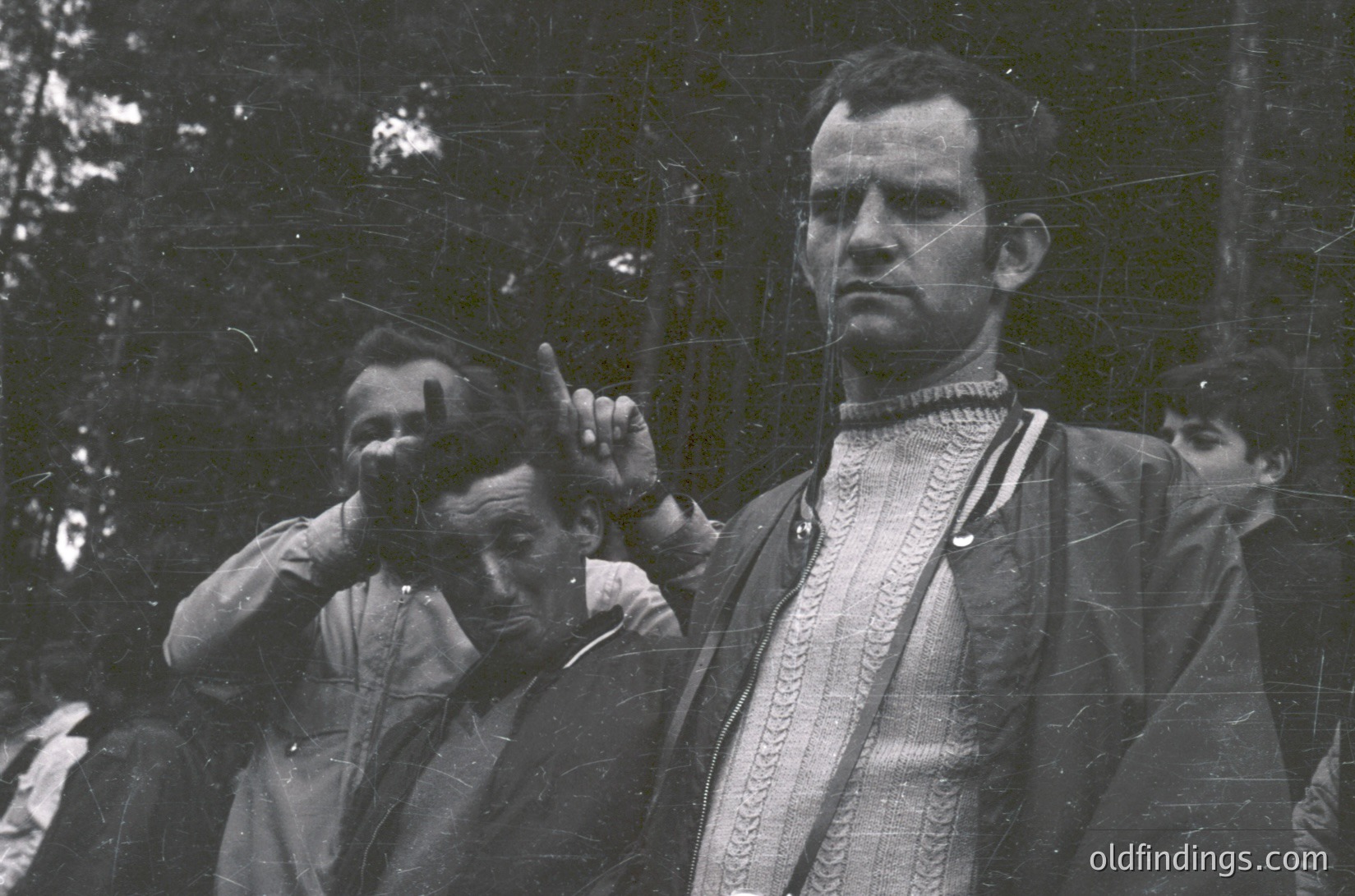 Vintage black-and-white portrait of two men in formal attire, likely from the early-to-mid 20th century. The man in focus wears a patterned vest and tie, adjusting his cap. Background suggests an outdoor setting with blurred greenery.