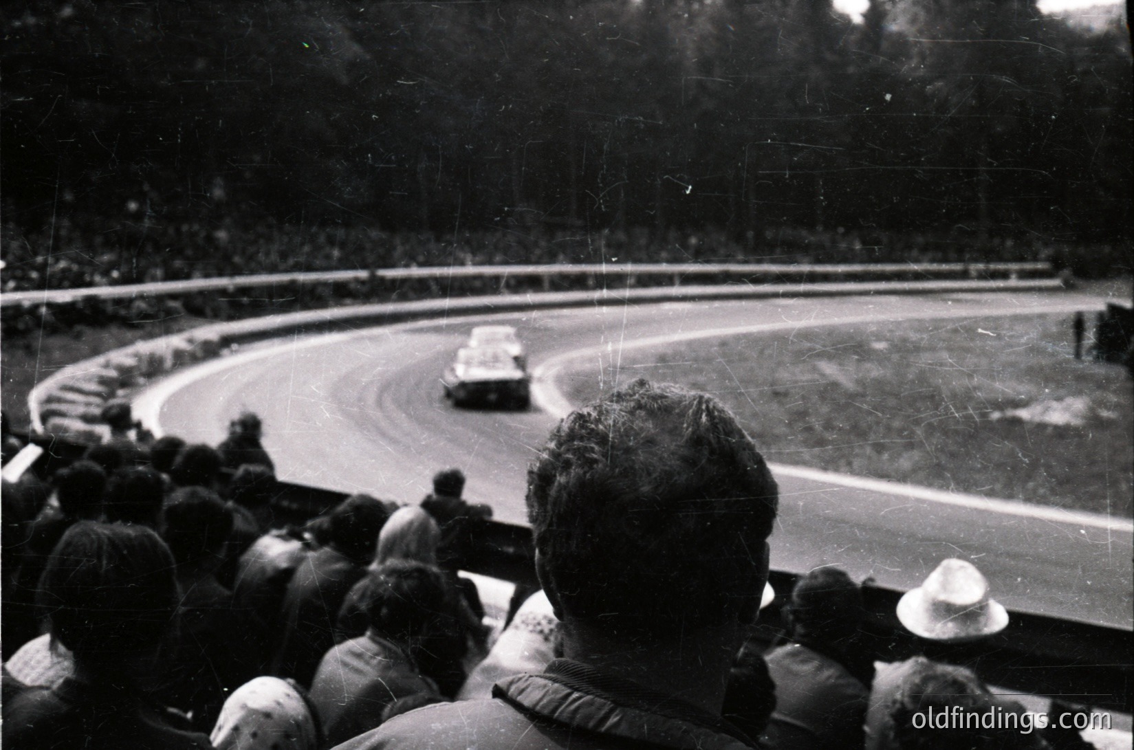 Black-and-white shot of a vintage race car navigating a banked oval track, surrounded by spectators. Mid-20th century racing atmosphere with blurred motion and dense crowd.