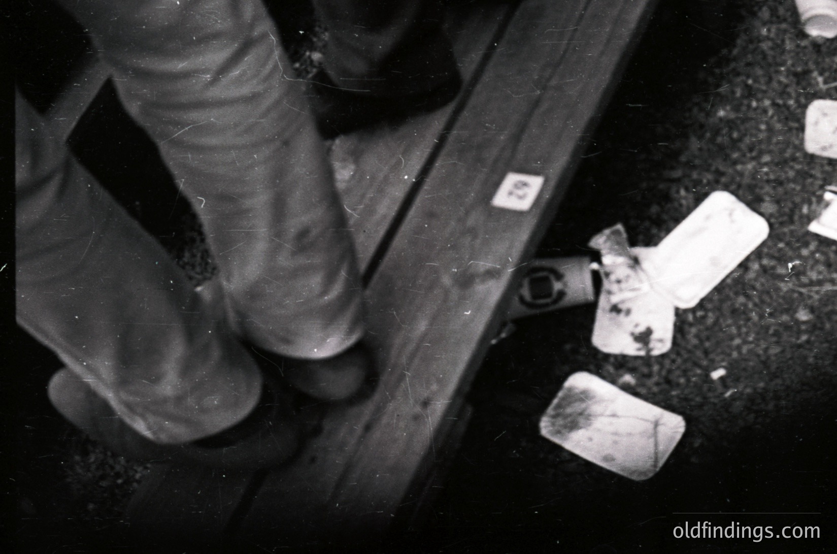 Black-and-white close-up of a person’s legs stepping over discarded soap bars on a metal rail. Industrial or institutional setting, likely a public restroom or laundry area. Soap bars appear worn, suggesting frequent use. Composition highlights hygiene infrastructure and utilitarian design.