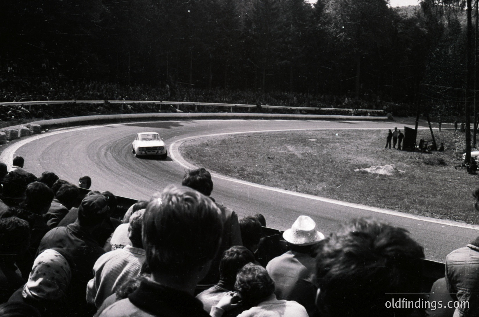Classic race scene: vintage sports car navigating a sharp curve on a tree-lined track, surrounded by spectators. Mid-20th century motorsport atmosphere with blurred motion emphasizing speed.