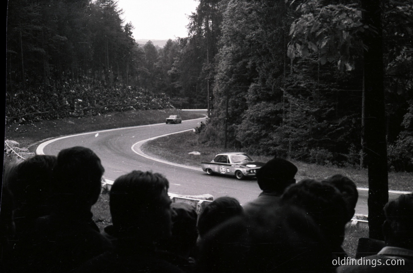 Classic black-and-white shot of a **rally race** on a winding mountain road, featuring vintage cars navigating tight curves. Spectators crowd the side, capturing the intensity of the event. Dense forest and misty hills frame the scene, suggesting a European alpine route. Likely mid-20th century (1950s–1970s) based on vehicle styling and film grain.