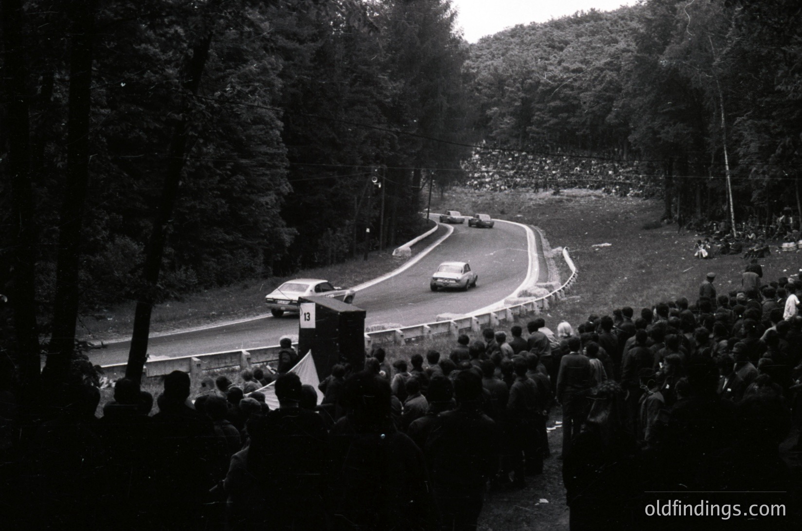 Vintage race car navigating a winding forest road during a rally, surrounded by dense spectators. Black-and-white photo captures mid-20th century motorsport excitement, likely or . Crowds line both sides of the road, emphasizing event scale.