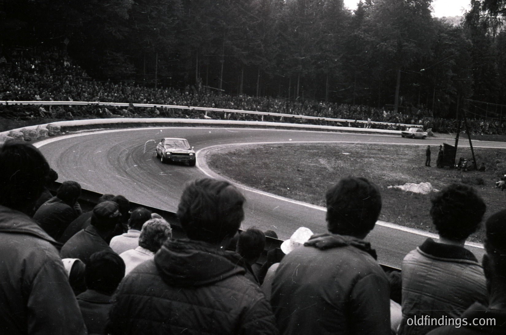 Black-and-white shot of a vintage race car navigating a sharp left curve on a tree-lined track, surrounded by dense crowds. Mid-20th century motorsport atmosphere with spectators in jackets, some holding cameras. Classic oval banking and gravel trap visible.