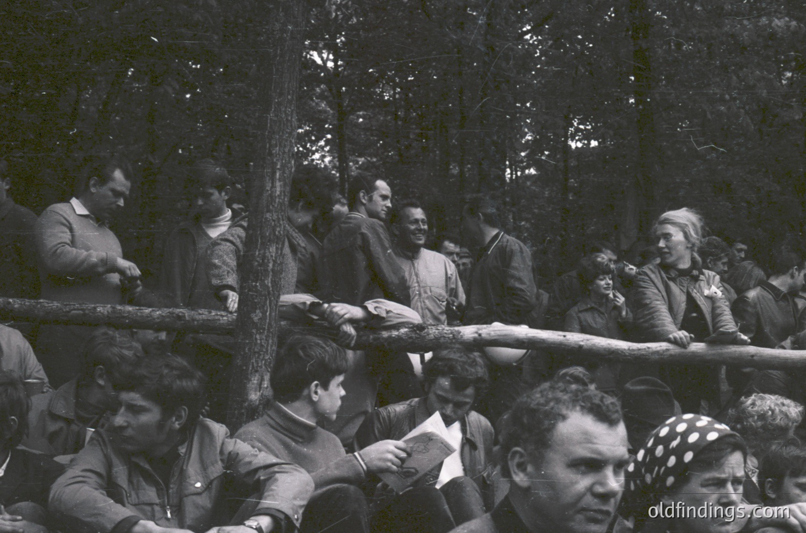 A black-and-white group photo of 1960s–70s outdoor gathering, likely a public event or rally. Crowd seated on wooden benches in a forested area, with dense trees framing the scene. Attendees wear mid-century formal/casual attire—jackets, ties, and headscarves. One man in foreground holds a folded paper, others engage in conversation. Atmosphere suggests a communal, possibly political or cultural event.