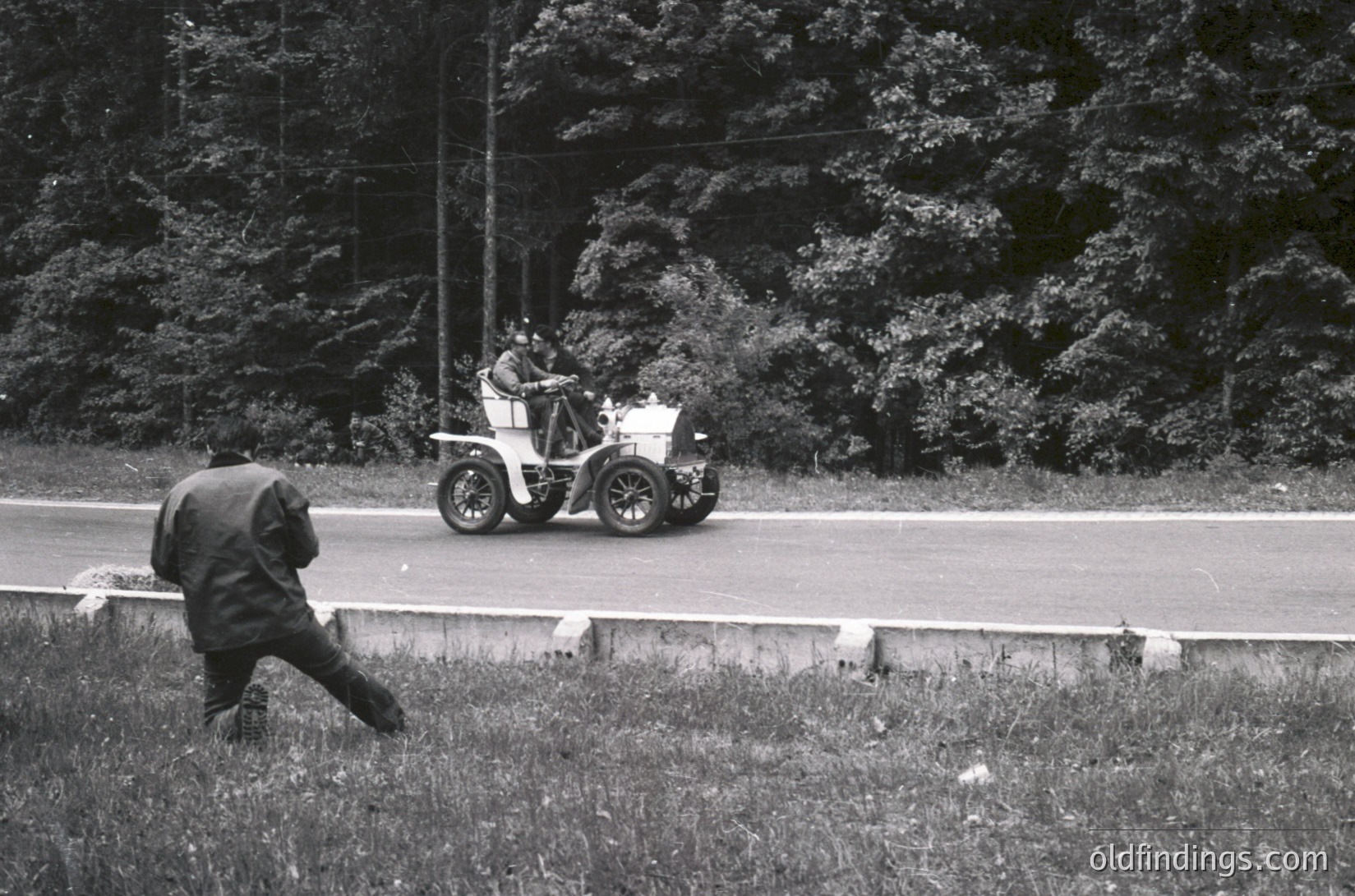 Vintage open-top vintage car racing on a rural road, with a lone figure in mid-stride beside the guardrail. Black-and-white shot captures motion and early automotive culture. Likely late 1920s–1930s, rural Europe/USA.