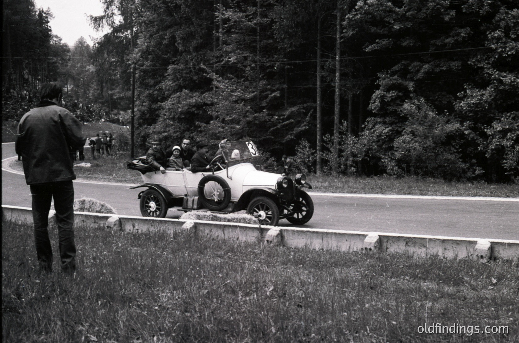 Vintage open-top race car on a rural road, surrounded by dense forest. Spectator in dark coat watches from guardrail. Mid-20th century roadside racing scene.
